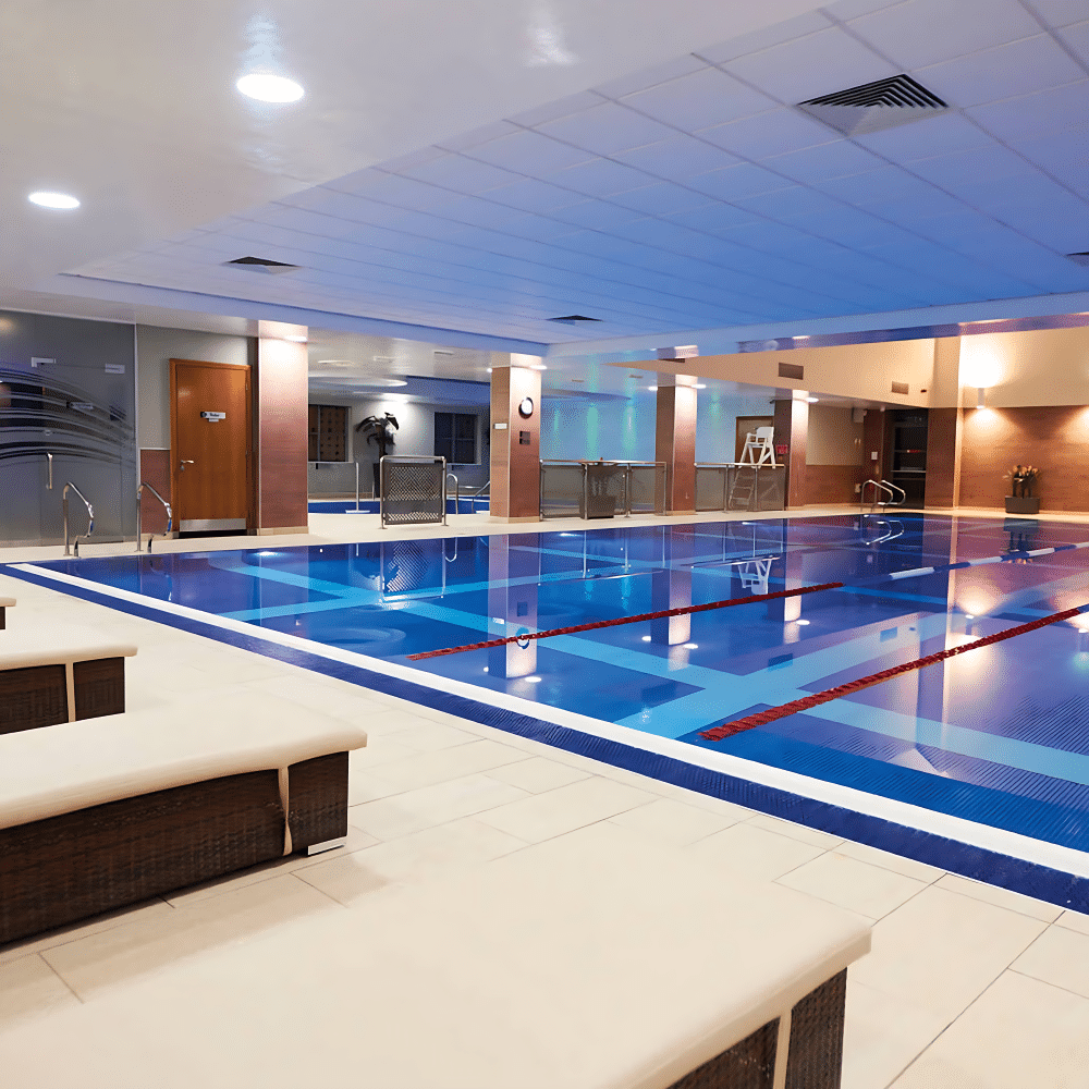 Indoor swimming pool with lounge chairs and lanes, under bright ceiling lights.