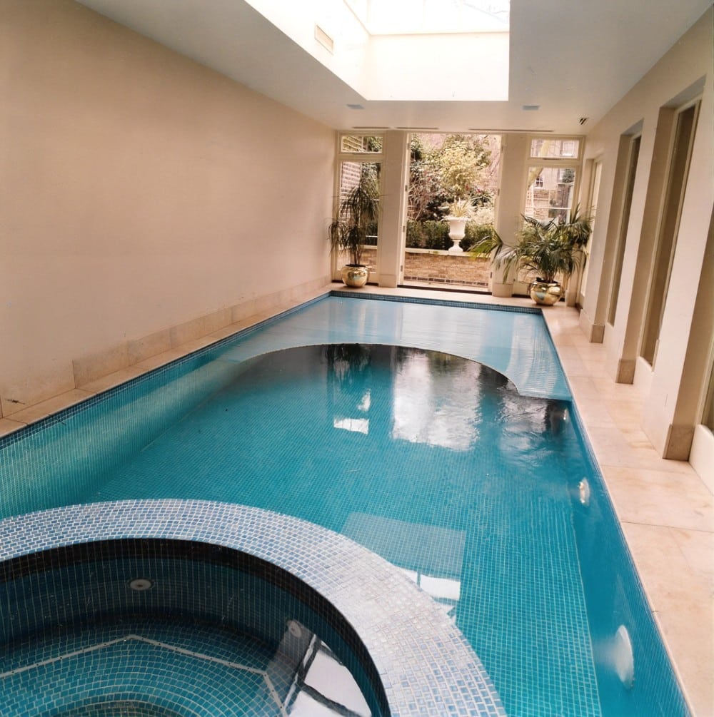 Indoor swimming pool with a hot tub, skylight, and plants near large windows overlooking a garden.