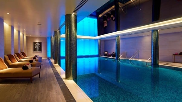 Indoor hotel pool with lounge chairs, blue lighting, and modern decor.
