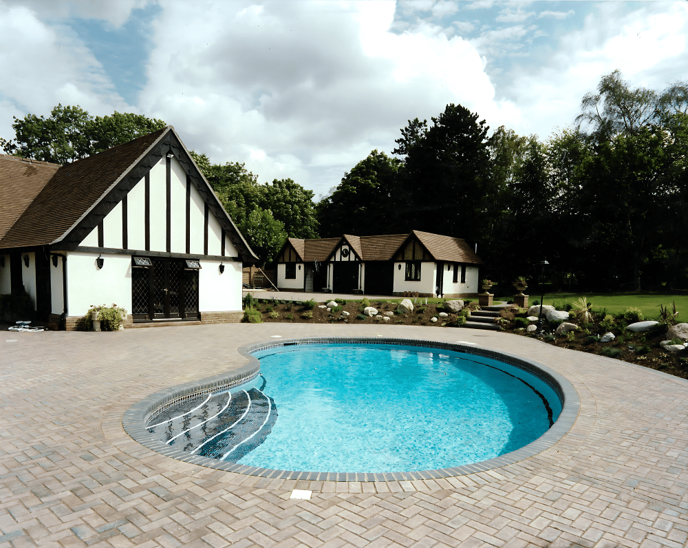 A kidney-shaped outdoor pool with brick paving beside a house and garden.